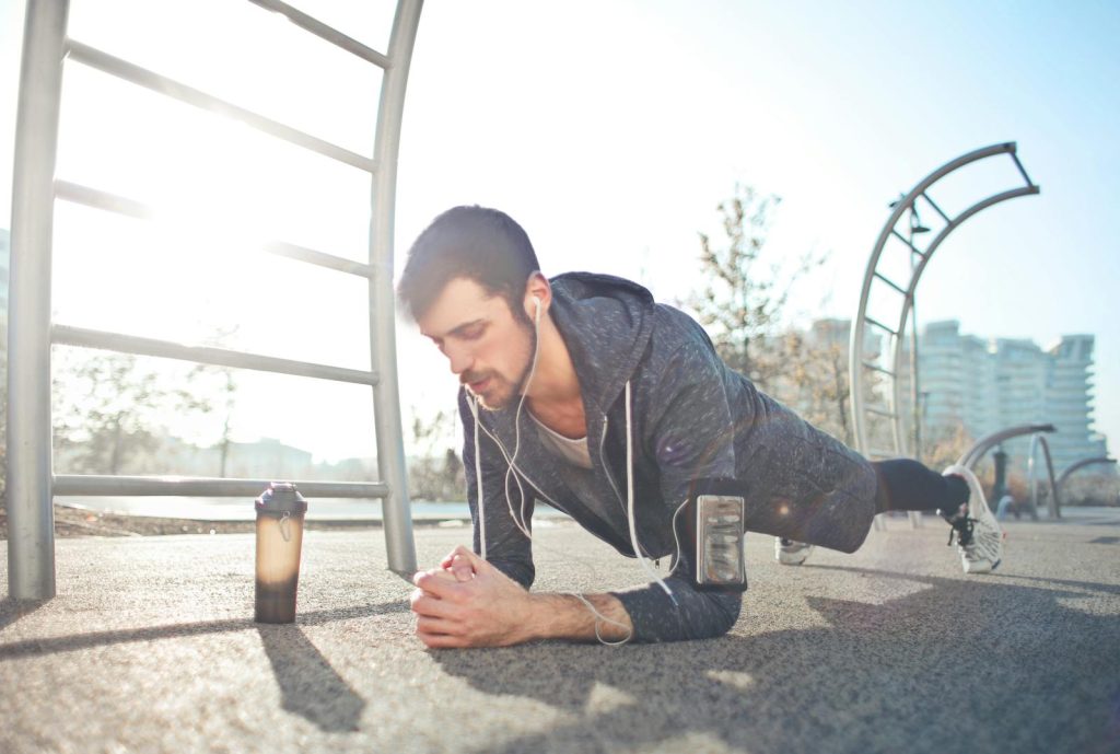 A young man performs a plank exercise outdoors with earphones, a phone, and a water bottle nearby.